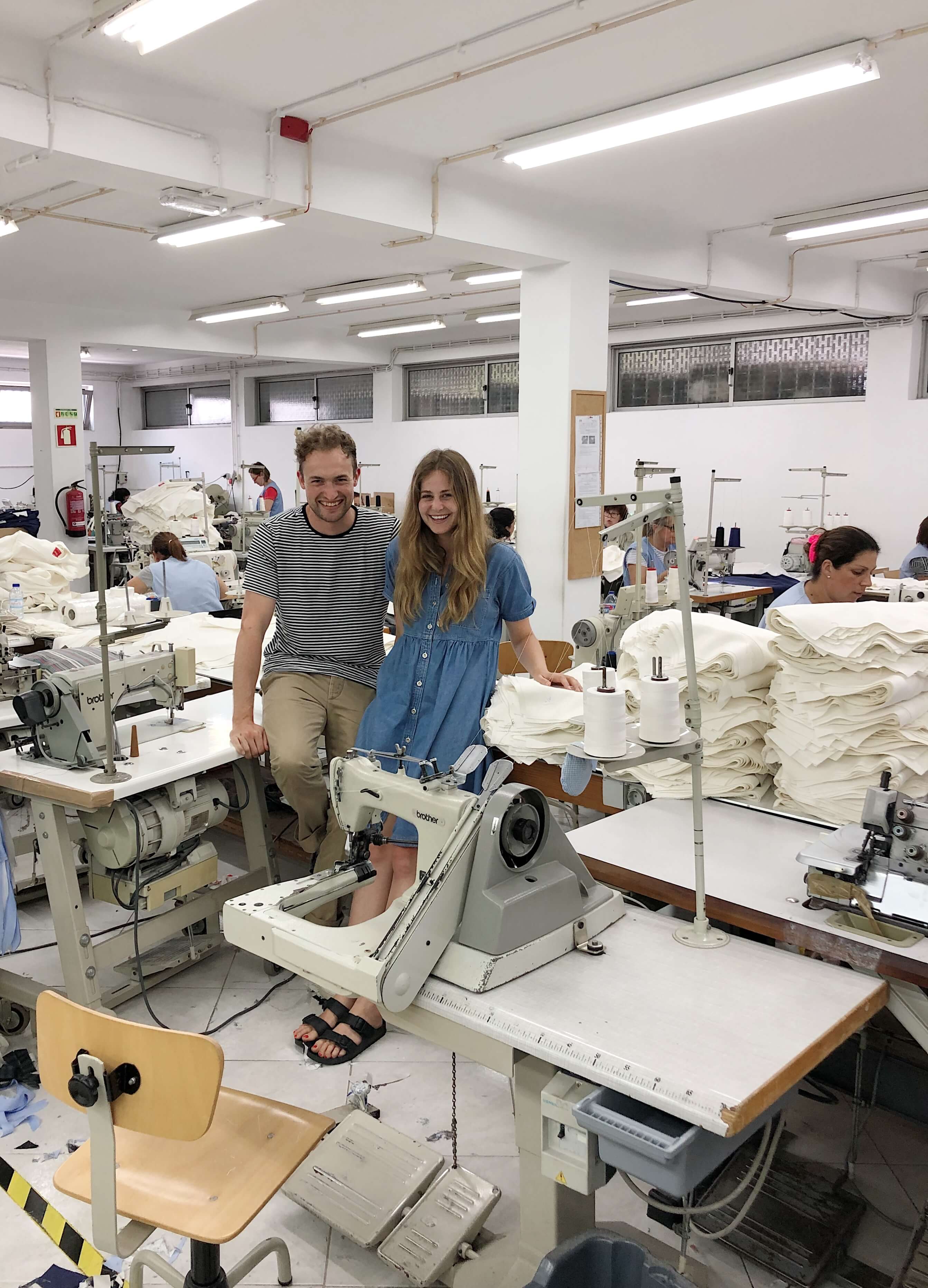 Huw and Becky standing at a clothing factory among workers sewing clothes