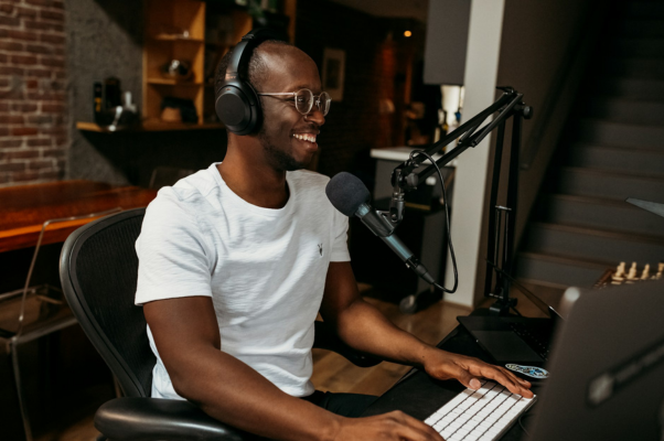 Man smiles at his computer screen while headphones with a microphone nearby to indicate creating youtube community posts. 