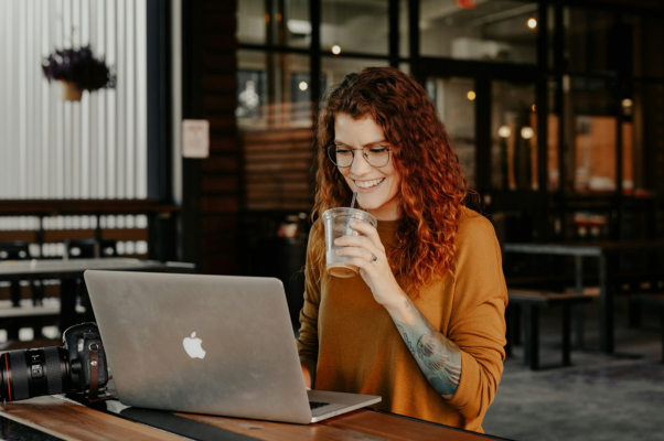 Woman working in a coffee shop sipping iced coffee while looking at her laptop to illustrate comparing Buffer vs. Hootsuite 