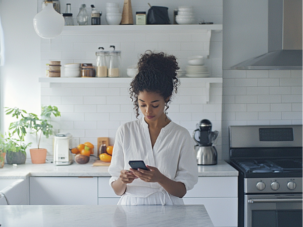 woman scrolling through her phone as she stands in a bright white kitchen to illustrate finding Instagram post ideas