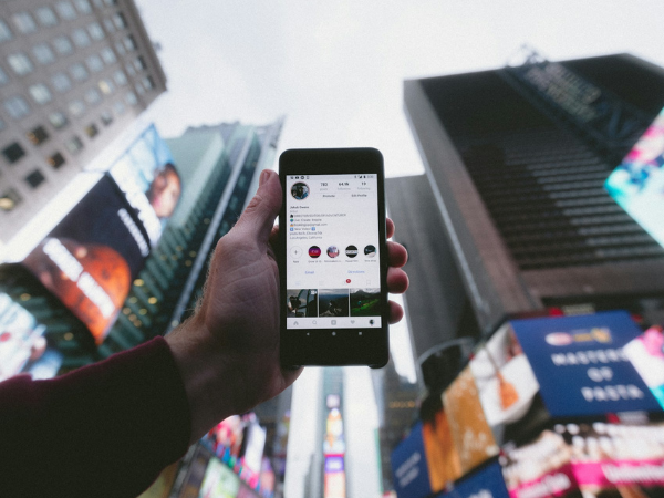 A man's hand holds a phone against the backdrop of several high-rise buildings to illustrate social media sites