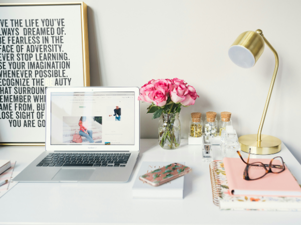 A macbook, phone, coloured notebooks, a gold lamp, and a vase of pink roses on a white desk