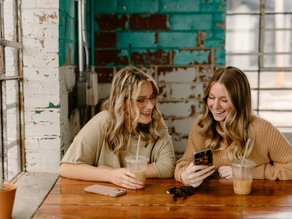 Two young women laughing as they look at a phone, while drinking iced coffee