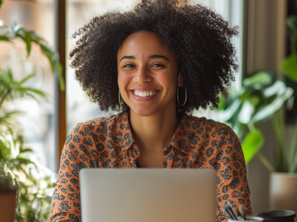 A young woman smiles as she sits in front of a laptop, to indicate becoming a creator