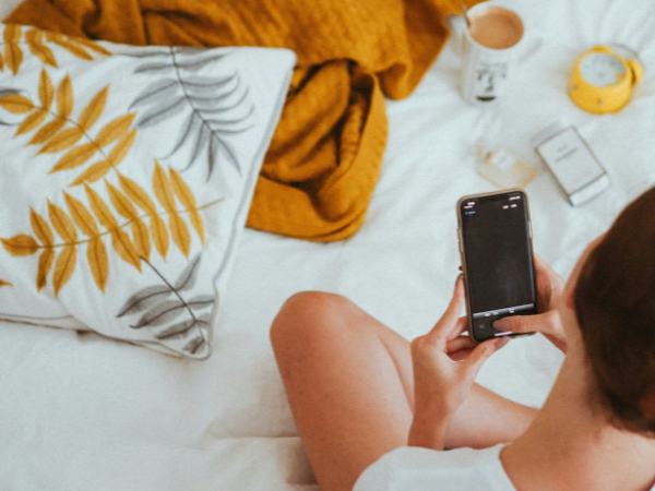 Woman sits on her bed while filming a video with her phone to illustrate using Instagram Trial Reels