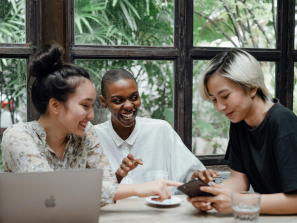 Three young women at a cofffe shop look at a laptop and phone to illustrate Social Media Engagement