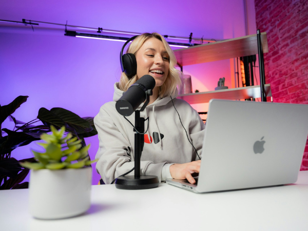 Smiling woman wearing headphones and hoodie records a podcast or YouTube video using a microphone and laptop in a modern setup with colorful lighting and a plant on the desk.