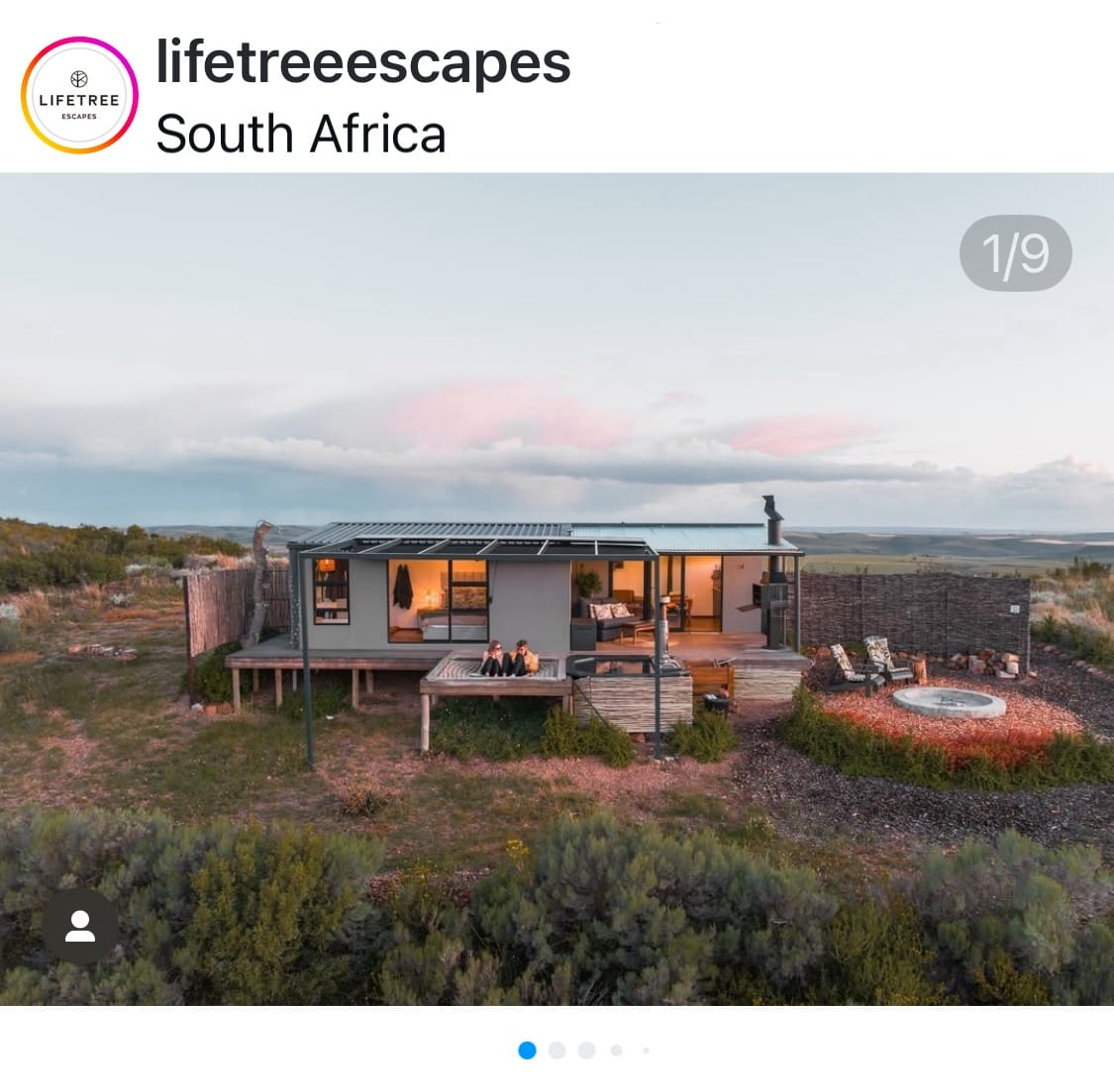 Alt text: Modern cabin in the South African countryside at sunset, with two people relaxing in an outdoor hot tub and a firepit area surrounded by natural vegetation.